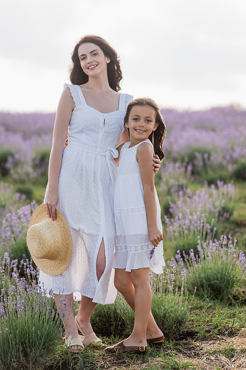 happy mother and daughter in white summer dresses smiling at camera in field