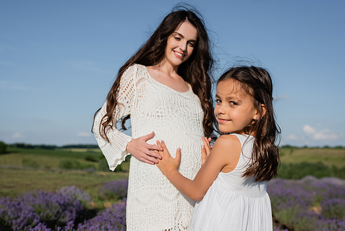 smiling girl embracing tummy of pregnant mother in openwork dress