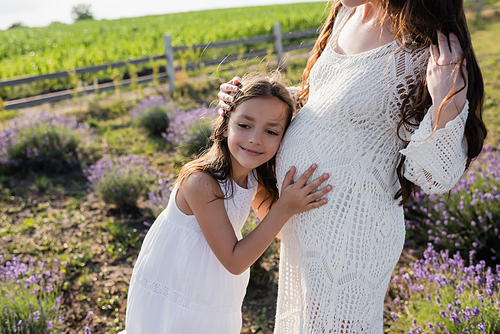 smiling child hugging belly of pregnant mother in blurred countryside field