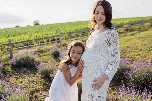 smiling kid embracing pregnant mom while standing in blurred meadow