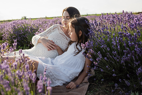 girl in white dress sitting near mother in field with blossoming lavender