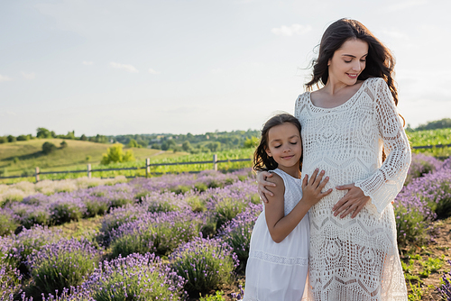 happy girl embracing tummy of pregnant mother in blooming lavender field