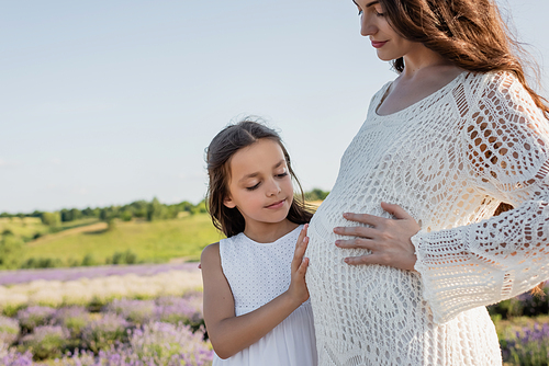 smiling girl hugging tummy of pregnant mother in blurred field