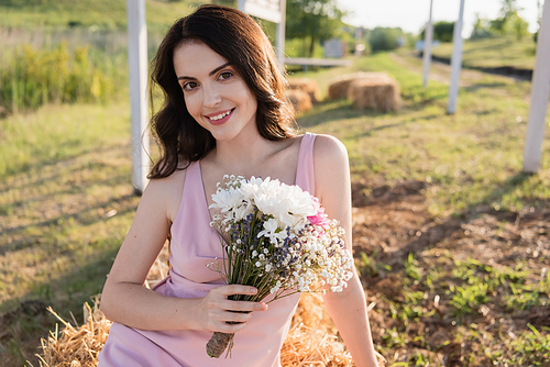 cheerful brunette woman with bouquet smiling at camera in countryside
