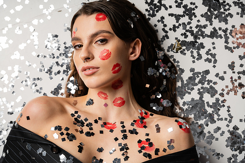 top view of brunette woman with red kiss prints looking at camera while lying near shiny confetti on grey background