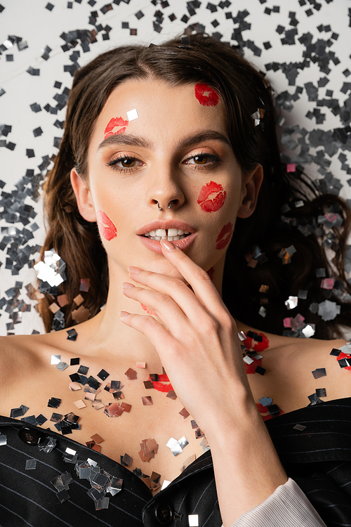 top view of sensual woman with makeup and red kiss prints touching lip near silver confetti on grey background
