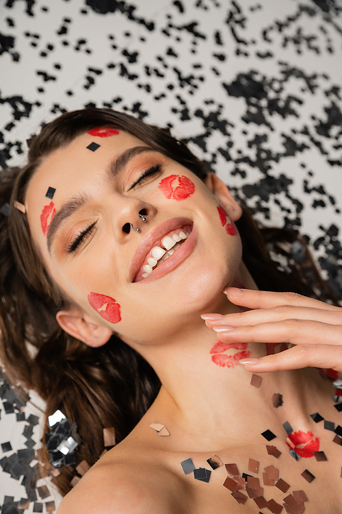 high angle view of cheerful woman with red lip prints and piercing lying with closed eyes near sparkling confetti on grey background