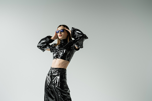 stylish young woman in black latex outfit and sunglasses posing isolated on grey