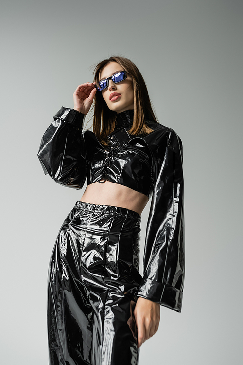 young woman in black latex clothing adjusting sunglasses isolated on grey