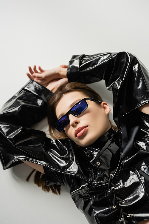 top view of brunette woman in black latex jacket and stylish sunglasses lying on grey