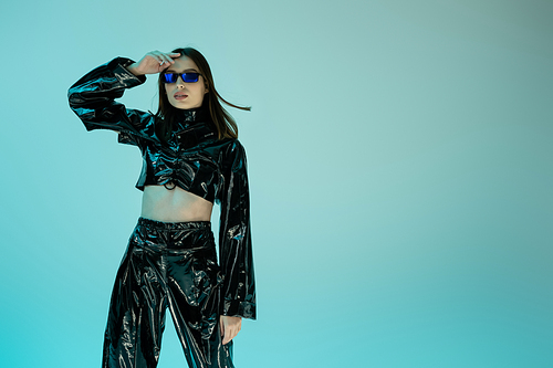 stylish young woman in black latex clothing and sunglasses standing isolated on blue