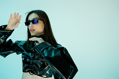 young woman in black latex clothing and sunglasses gesturing isolated on blue background