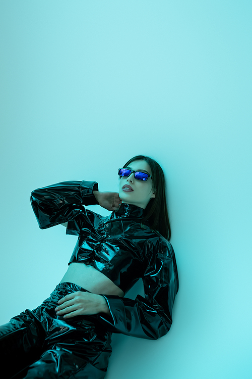 stylish young woman in sunglasses and black latex clothing sitting on blue background