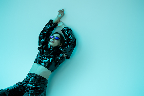 stylish young woman in sunglasses and black latex clothing posing with raised hands on blue background