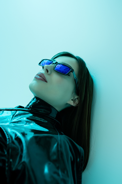 portrait of young woman in sunglasses and black latex clothing posing on blue