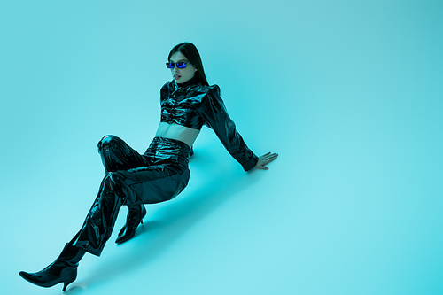 full length of stylish young woman in latex clothing and high heels sitting on blue background