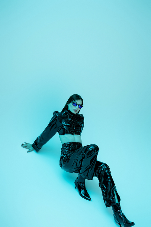full length of young woman in sexy latex clothing and high heels sitting on blue background
