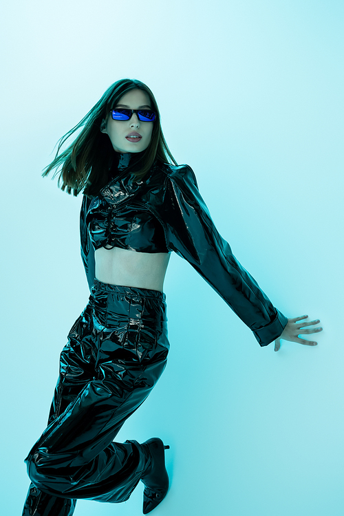 brunette woman in sunglasses and sexy latex clothing posing on blue background