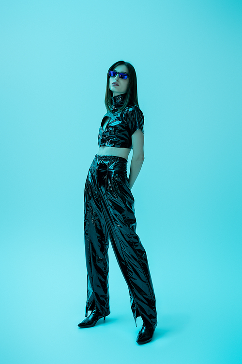full length of stylish young woman in latex crop top and trousers standing on blue