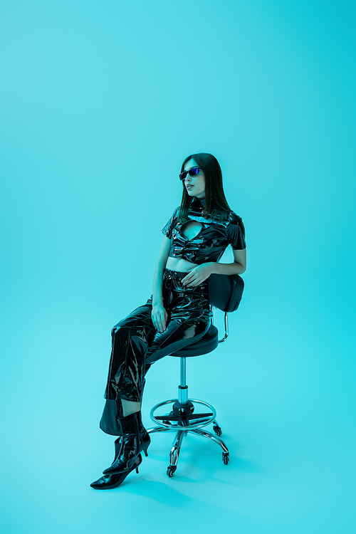 full length of brunette woman in latex crop top and trousers sitting on stool while looking away on blue