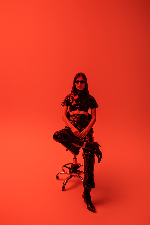 full length of brunette woman in latex crop top and trousers sitting on stool while looking at camera on red