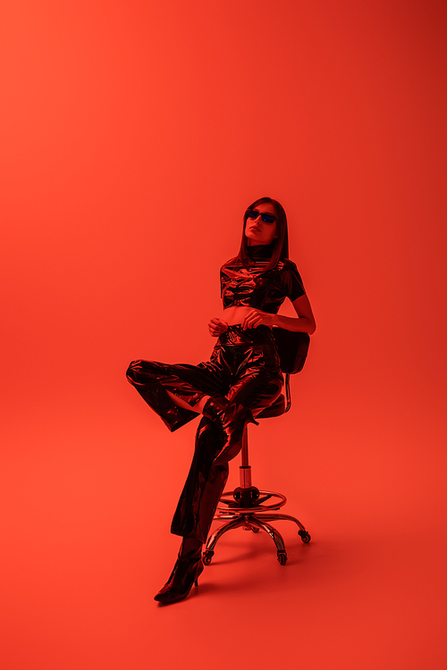 full length of stylish woman in latex crop top and trousers sitting on stool while looking at camera on red