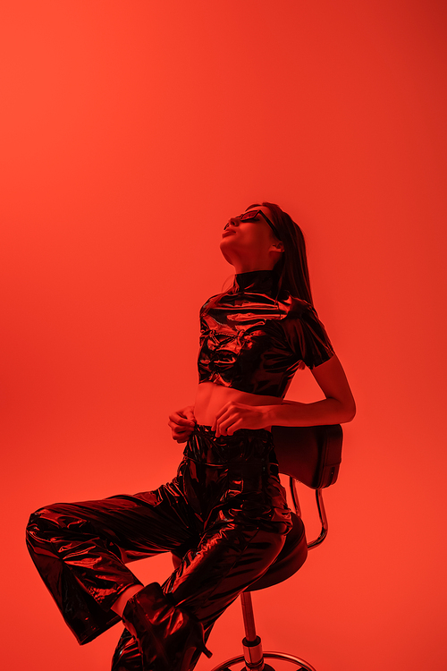 stylish woman in latex crop top and trousers sitting on stool isolated on red