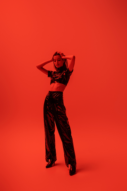 full length of stylish woman in latex crop top and trousers posing with hands on head on red