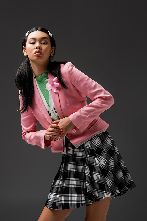 Pretty asian woman in skirt and pink jacket looking at camera isolated on grey