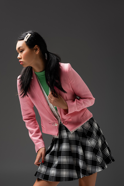 Stylish asian model in checkered skirt and pink jacket posing isolated on grey