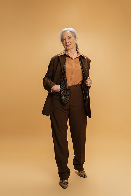 full length of senior woman in brown formal wear standing with hand in pocket on beige background