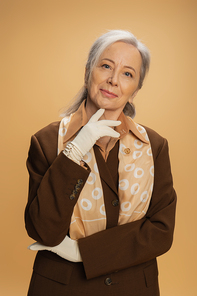portrait of pleased senior woman in white gloves and brown suit looking at camera isolated on beige