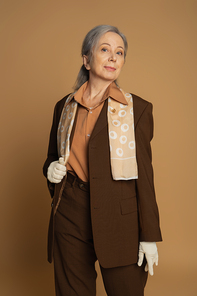 senior woman in brown formal wear with scarf and white gloves looking at camera on beige