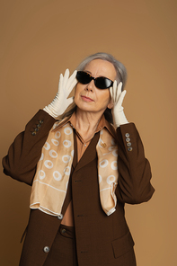 senior woman in brown formal wear and white gloves adjusting sunglasses isolated on beige