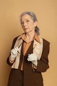 senior woman in brown formal wear and white gloves holding sunglasses isolated on beige
