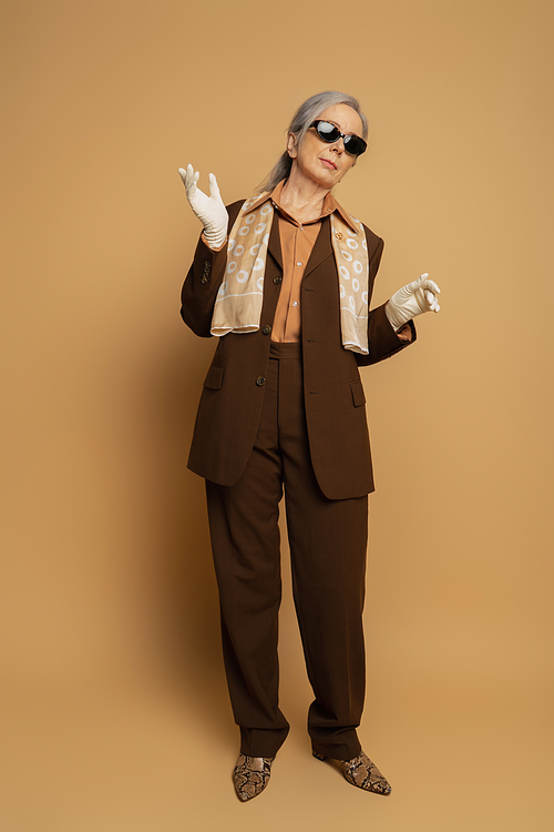 full length of senior woman in brown formal wear and white gloves standing on beige