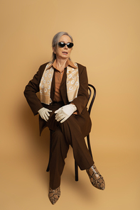 full length of senior woman in brown formal wear and sunglasses sitting on wooden chair on beige