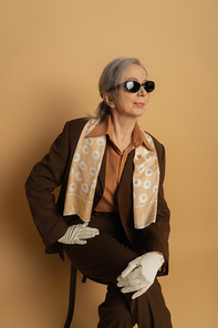 senior woman in brown suit and sunglasses sitting on wooden chair on beige