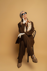 full length of thoughtful senior woman in brown formal wear and sunglasses sitting on wooden chair on beige