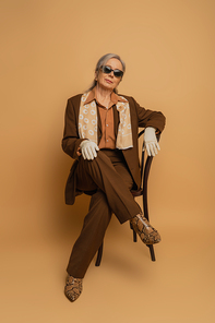 full length of elderly woman in brown formal wear and sunglasses sitting on chair and looking at camera on beige