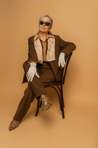 full length of elderly woman in brown suit and gloves sitting on chair and looking at camera on beige