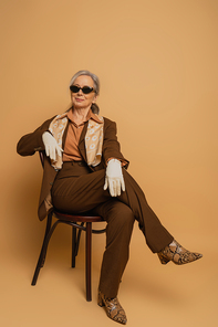 full length of cheerful elderly woman in brown suit and sunglasses sitting on chair and looking at camera on beige
