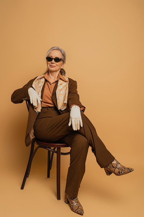 full length of cheerful elderly woman in brown suit and sunglasses sitting on chair and looking at camera on beige