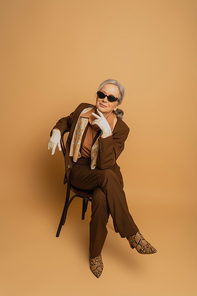 full length of smiling elderly woman in brown suit and sunglasses sitting on chair and looking at camera on beige