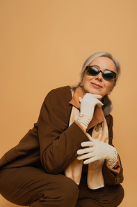 smiling and stylish senior woman in sunglasses and suit sitting isolated on beige