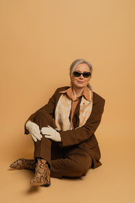 full length of cheerful senior woman in sunglasses and suit sitting and smiling on beige background