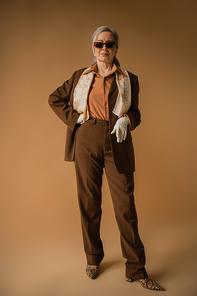 full length of senior woman in brown suit and white gloves posing with hand on hip on beige