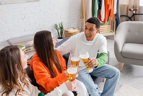 cheerful women and amazed african american man clinking glasses of beer in living room