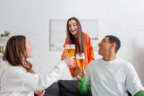 positive women and amazed african american man clinking glasses of beer in living room