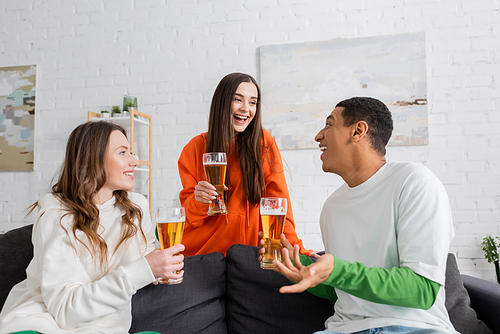 positive women and happy african american man holding glasses of beer in living room